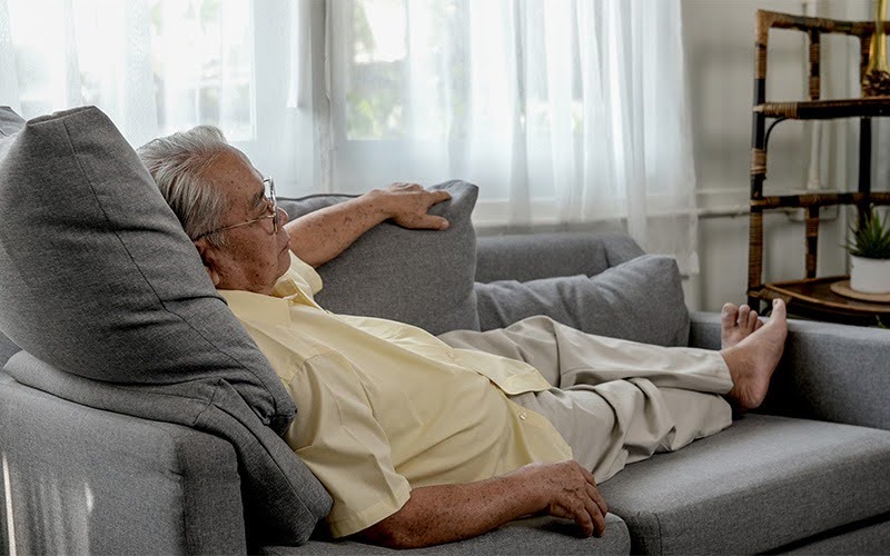 Older adult relaxes on gray sectional sofa at home.