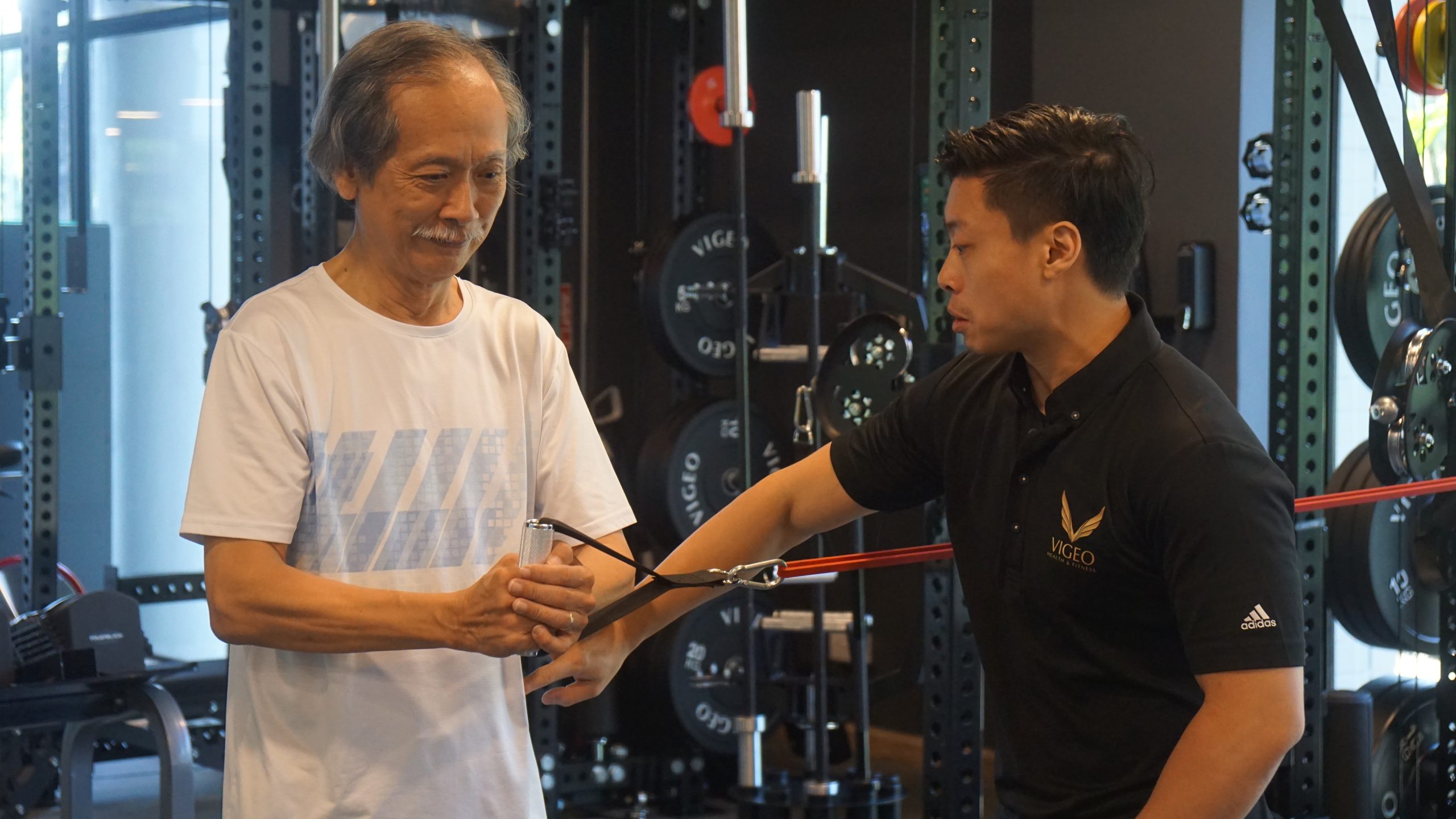 Vigeo trainer guiding older man through cable resistance exercise.